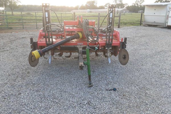 Red farm equipment with hydraulic hoses and wheels on gravel surface.