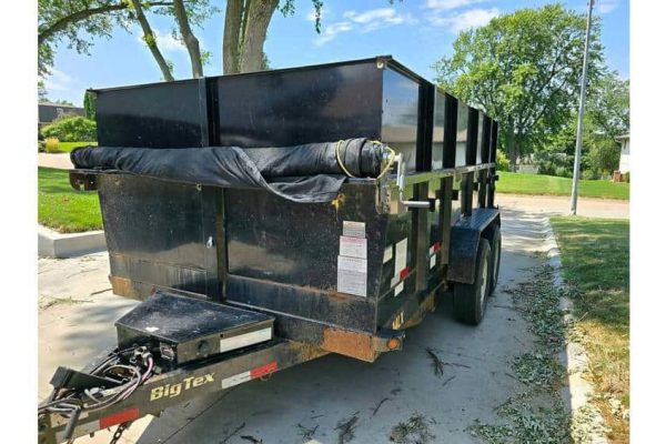 Heavy-duty black dump trailer with dual axles, parked on a paved surface in a green outdoor setting.