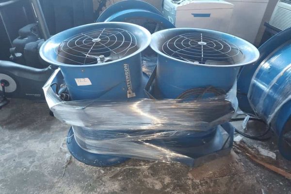 Two blue industrial air circulators with protective grilles, wrapped in plastic, on a concrete floor.