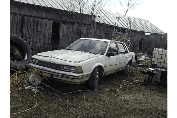 Old beige 1980s sedan parked in a rustic yard with wooden shed, tires, and equipment around.
