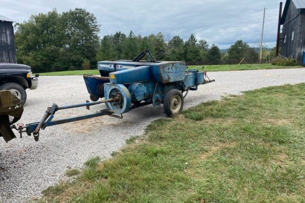 Old blue agricultural seeder machine with rust and wear, on a gravel surface with trees and buildings in background.