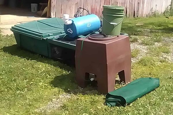 Assorted outdoor storage bins, buckets, and a rolled-up tarp on grass near a wooden shed.