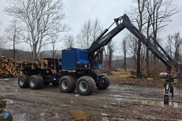 Forestry grapple loader with log pile in muddy outdoor setting.