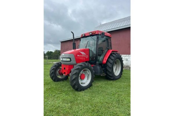 Red McCormick tractor with enclosed cab, parked on grass near a red barn under cloudy sky.