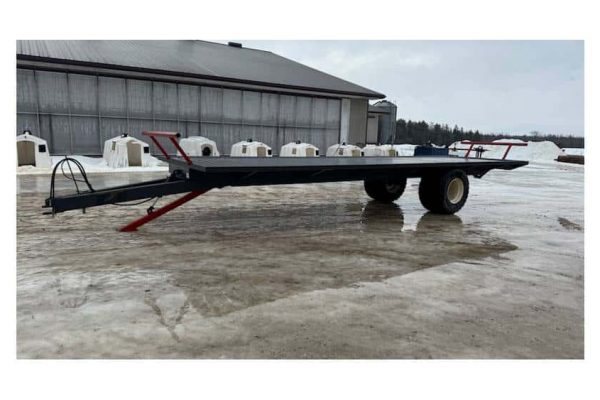 Flatbed trailer with dual axles parked outdoors on a snowy surface near a large barn.