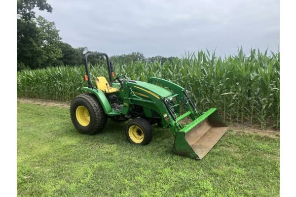 A.LT tag: Green John Deere tractor with front loader attachment on grassy field near cornfield, under cloudy sky.