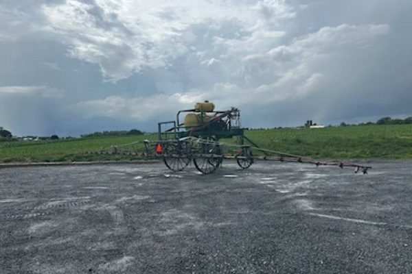Farm sprayer on wheeled cart with tank and spray booms under cloudy sky.