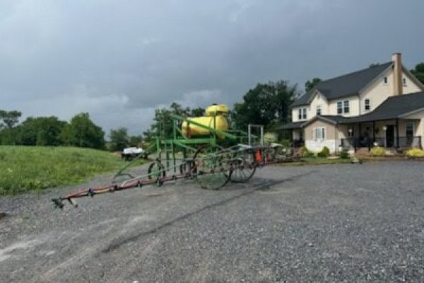 Agricultural sprayer with yellow tank and green frame on gravel driveway near house.