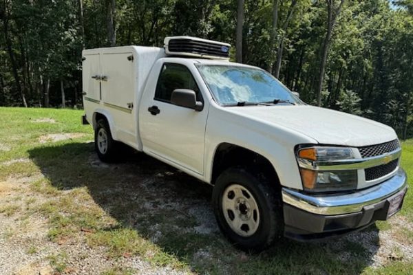 White Chevrolet Colorado utility truck with refrigeration unit on a grassy area.