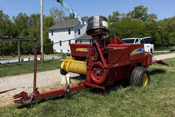 Red hay baler machine with yellow pickup and black engine, outdoors on green grass under clear blue sky.