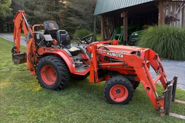 Kubota L Series compact tractor with backhoe attachment, orange color, in a grassy yard with barn and trees.