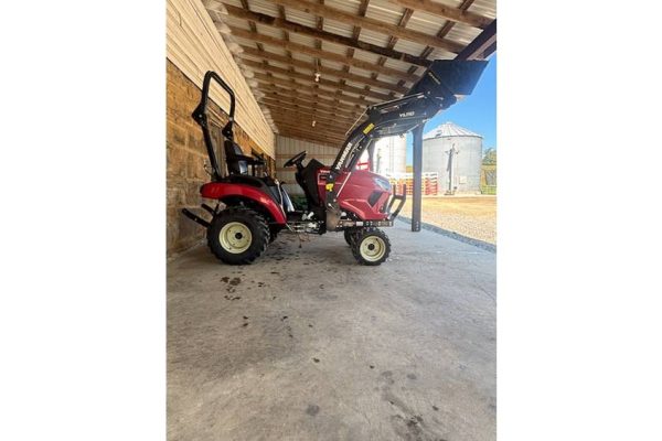 Red compact tractor with front loader attachment parked under a wooden shelter.