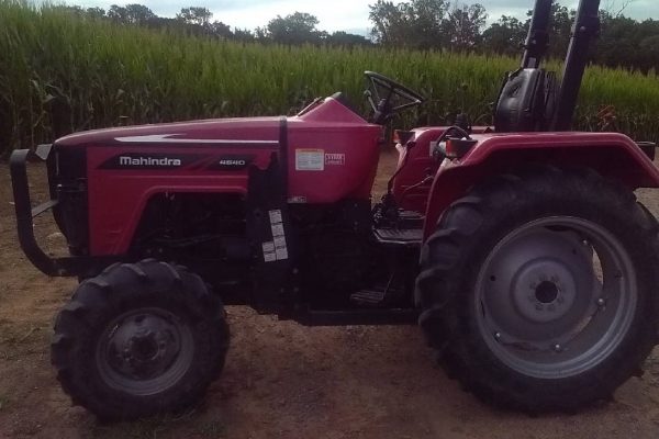 Mahindra tractor from 2020 model year, red color, parked on farm field with green crops and cloudy sky in background.