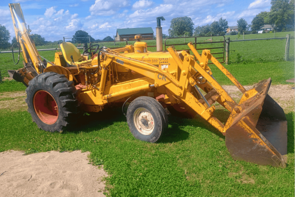 Yellow Case 580 backhoe loader with front bucket and rear excavator arm on grassy field.