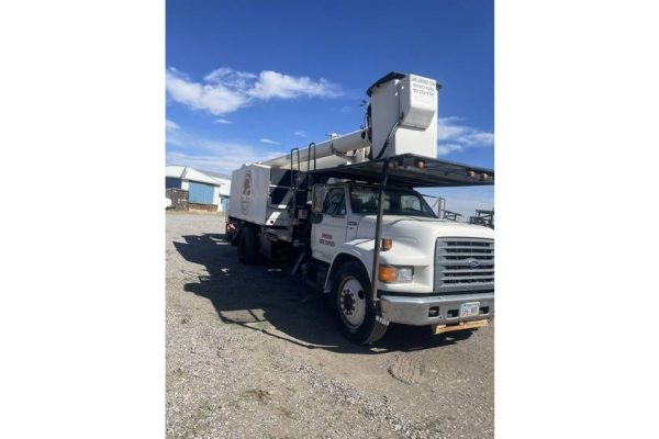 White bucket truck with aerial lift, likely from 2000s, parked outdoors on gravel under blue sky.