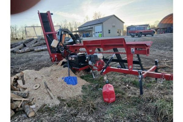 Red log splitter machine on a farm with a house and trees in the background during daytime.
