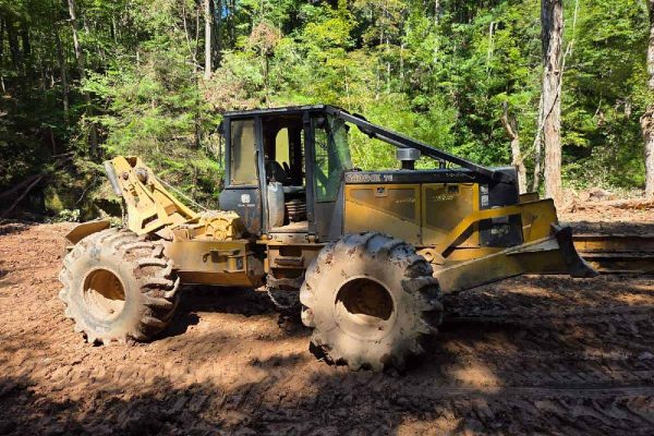 Yellow Caterpillar skid steer loader with enclosed cab on a dirt site surrounded by trees.