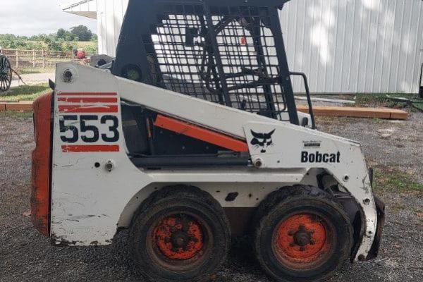 Bobcat 553 skid steer loader with enclosed cab and orange wheels, parked outdoors.