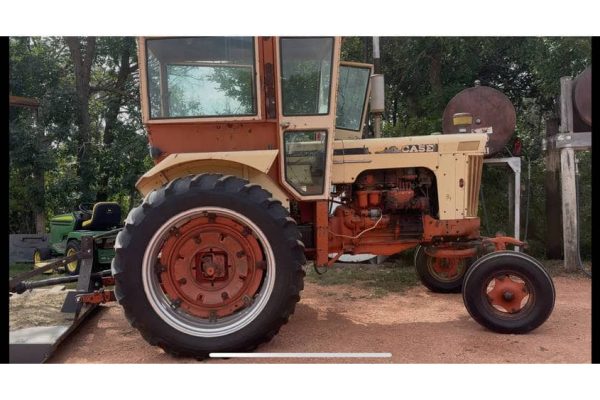 Vintage 1960s beige tractor with large rear tires and enclosed cab, parked outdoors among trees.
